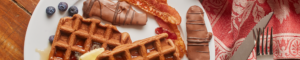 Plate of muddy pigs chocolate covered bacon, waffles, blueberries and utensils.